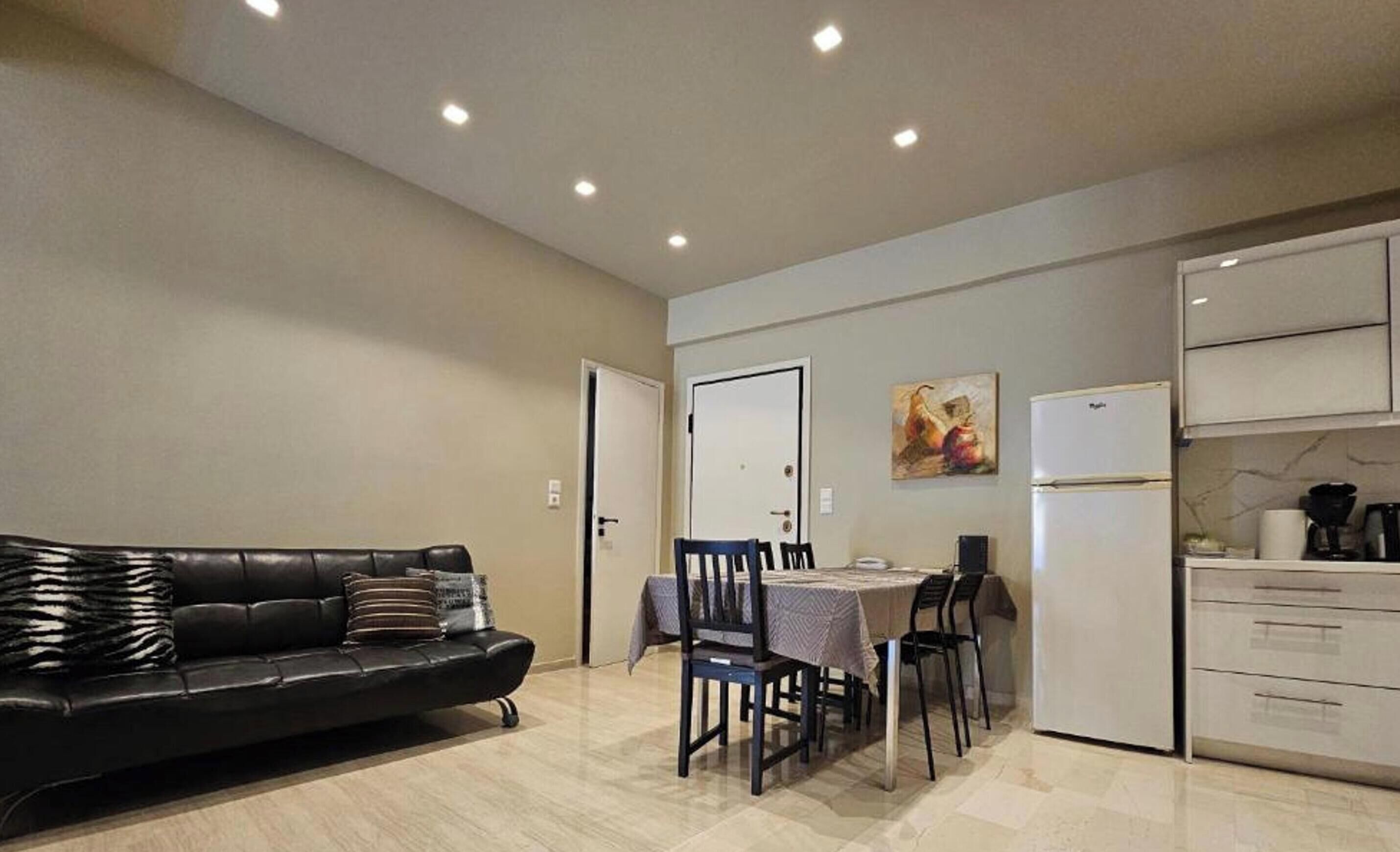 Ein Wohnzimmer mit schwarzem Ledersofa, einem Esstisch mit Stühlen, einer Küchenzeile mit Schränken und einem Kühlschrank, sowie einer Tür zu einem weiteren Raum.