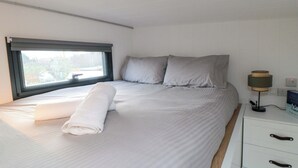 Ein ordentlich bezogenes Bett mit Kissen, einer Nachttischlampe und einem Fenster mit Jalousien.