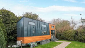 Ein modernes, kompaktes Haus mit einem hölzernen Unterteil und einem dunklen Oberteil, mit einer kleinen Veranda und einem Kiesweg zum Eingang.