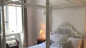 En seng med himmelseng, hvid baldakin, trægavl og et natbord med en lampe.