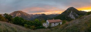 Paisaje rural con una casa en una colina, rodeada de montañas y vegetación.