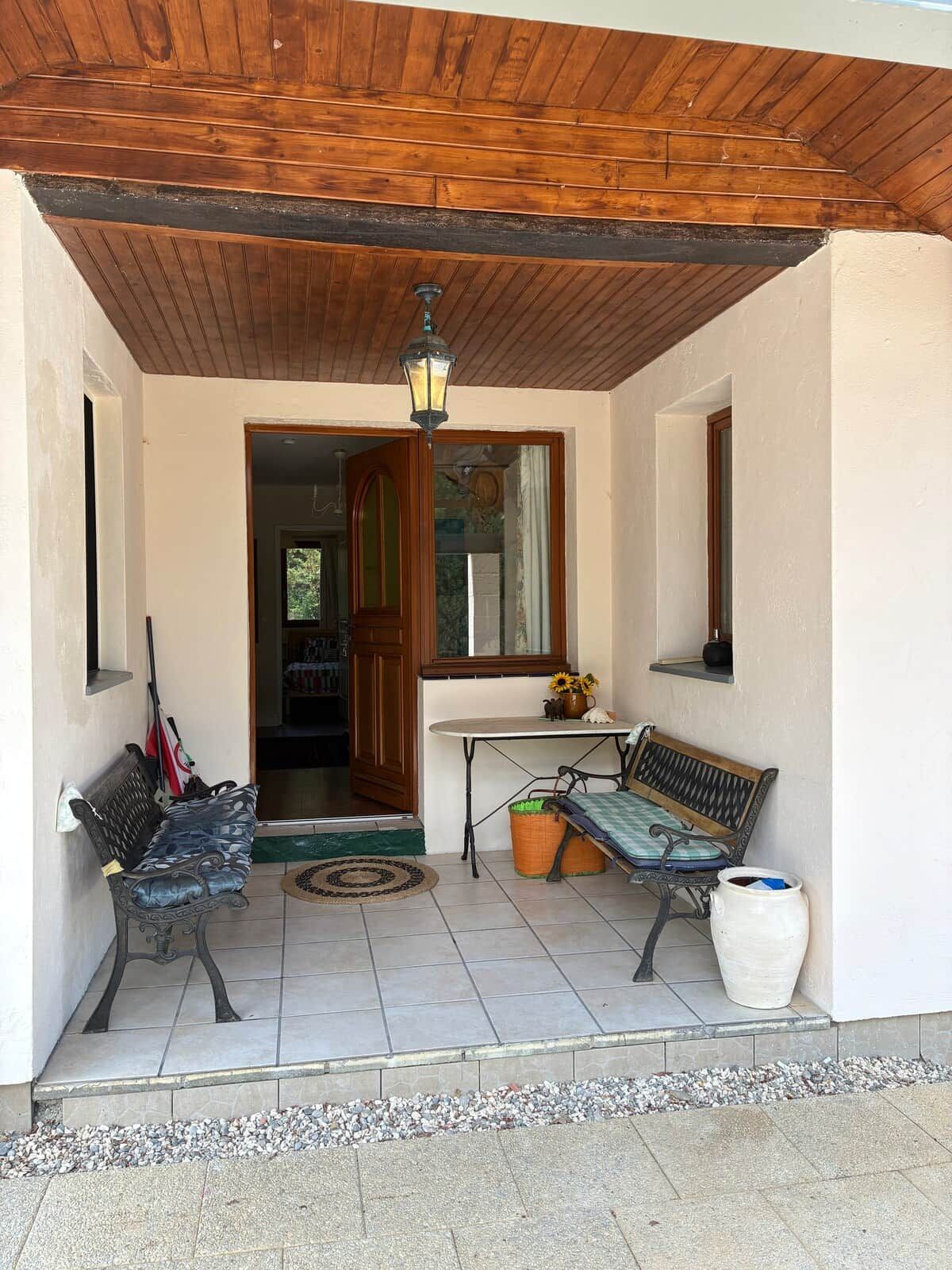 A covered porch with a wooden ceiling, a bench, a table, a potted plant, and a door leading inside.