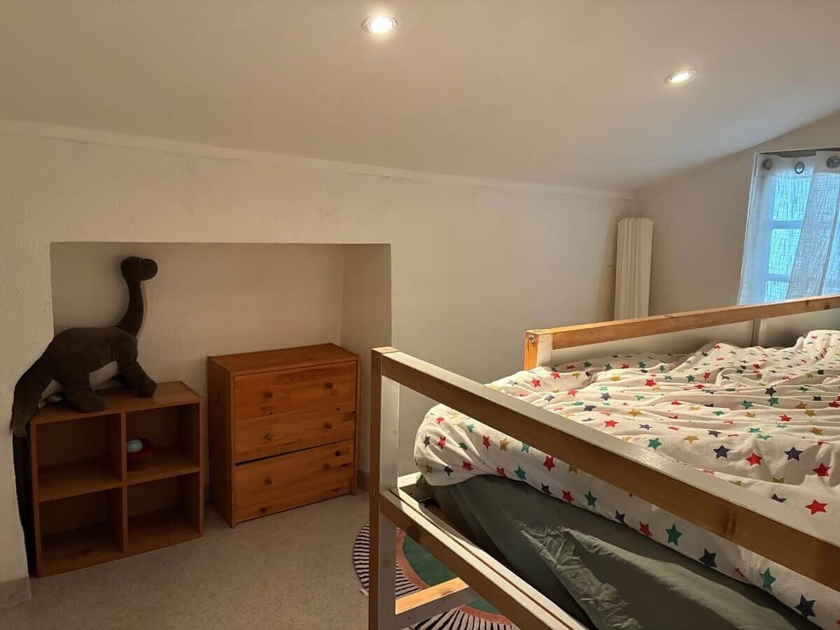 Une chambre avec un lit superposé, une commode en bois, une étagère avec un chat et une fenêtre avec des rideaux.