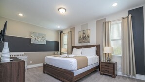 Une chambre à coucher comprenant un lit, des tables de chevet, une commode, des œuvres d’art accrochées aux murs et une fenêtre avec des rideaux.