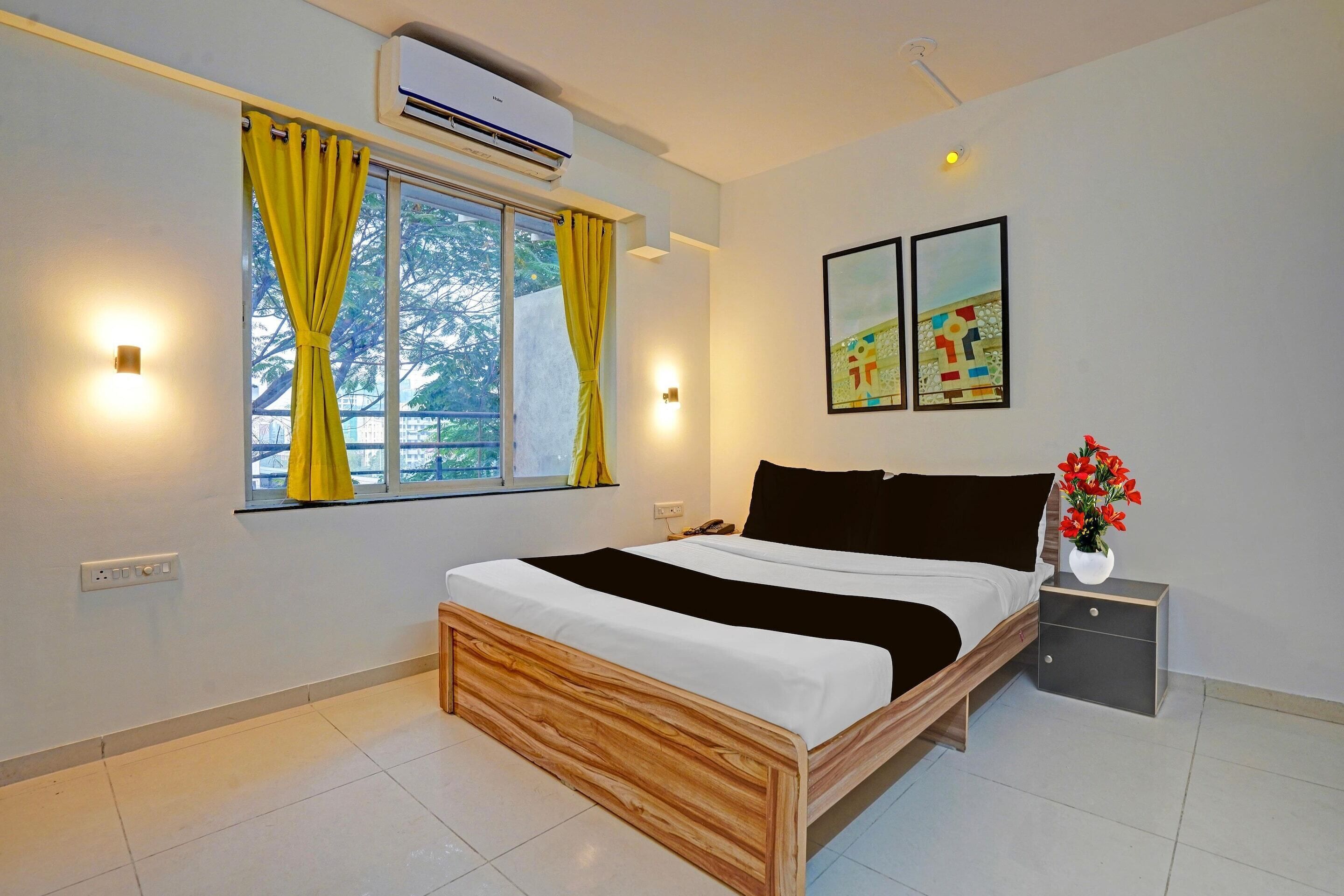 A bedroom with a bed, a nightstand, a window with curtains, wall-mounted lights, and two framed pictures.