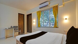 A hotel room with a bed, a desk, a chair, an air conditioner, and a window with curtains. - Classic With Balcony I- Siri Lodge (Pune)