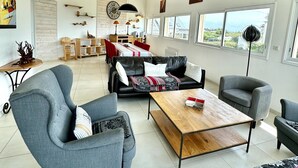 A spacious living area with a sofa set, a coffee table, and a dining table. There are multiple windows providing natural light and views. - Neiz Vran House Facing the Sea (Kerlouan)