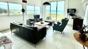 A spacious living area with a sofa, chairs, a coffee table, and a desk with a television. Large windows offer a view of the outdoors. - Neiz Vran House Facing the Sea (Kerlouan)