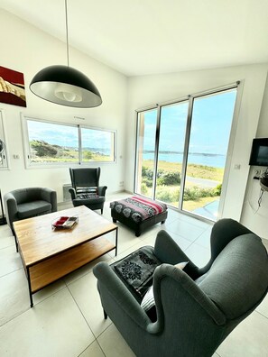 A modern living room with a large window, a sofa, a coffee table, and a television. - Neiz Vran House Facing the Sea (Kerlouan)