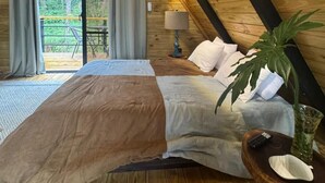 A cozy wooden cabin bedroom with a bed, a nightstand, a lamp, and a window overlooking a forest. - Alpine Villa | Mountain| Fire pit| Heated Jacuzzi (Jarabacoa)