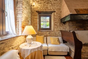 Ein Zimmer mit Steinwänden, einem Sofa, einem Tisch mit Lampe und einem Fenster mit Blick auf Grünflächen.