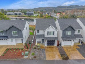 A row of modern houses with attached garages, a landscaped front yard, and a view of a lake and hills in the background. - Riverview Cottage– Walk to Beach, Views Everywhere! (Seaside)