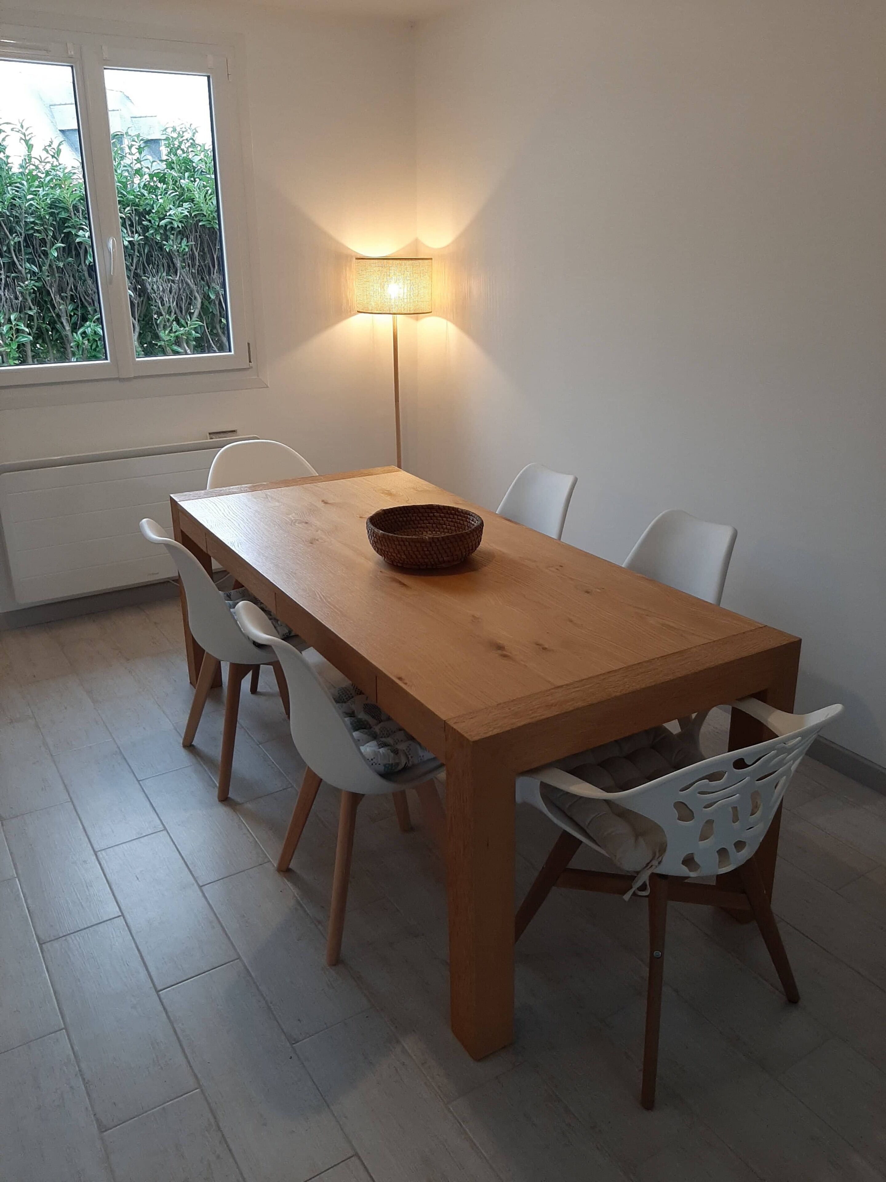 A dining area with a wooden table, four white chairs, a woven bowl, and a floor lamp.