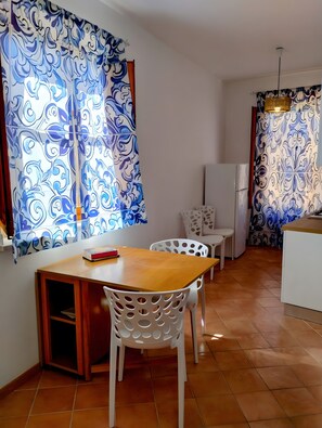 A compact kitchen with a dining area, white chairs, a wooden table, and patterned curtains. - Apartment 'Casa Ariele' with Mountain View, Private Terrace and Air Conditioning (San Vito Lo Capo)