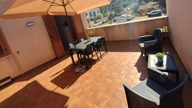 A balcony with a table and chairs, a large umbrella, and a view of a hilly landscape. - Apartment 'Casa Ariele' with Mountain View, Private Terrace and Air Conditioning (San Vito Lo Capo)