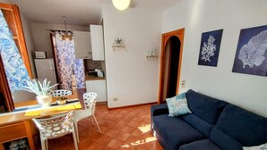 A living room with a sofa, a dining table with chairs, a kitchen area, and a hallway. - Apartment 'Casa Ariele' with Mountain View, Private Terrace and Air Conditioning (San Vito Lo Capo)