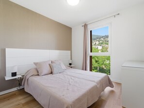 A bedroom with a bed, a nightstand, a lamp, a window with a view of buildings, and a ceiling light. - Charming apartment for 4 people with a balcony (Grasse)