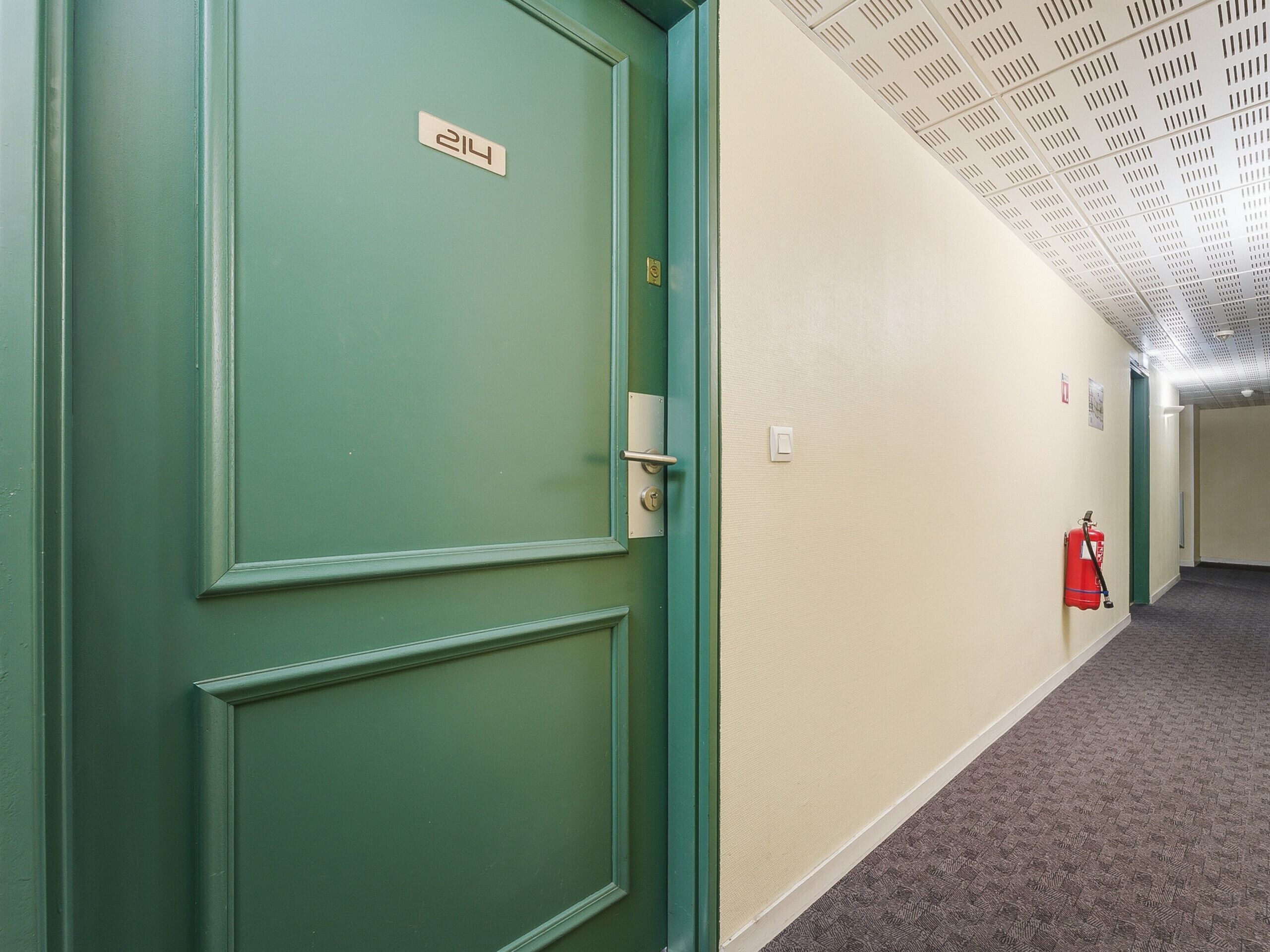 A hallway with a green door marked '214', a fire extinguisher on the wall, and a ceiling with a grid pattern.
