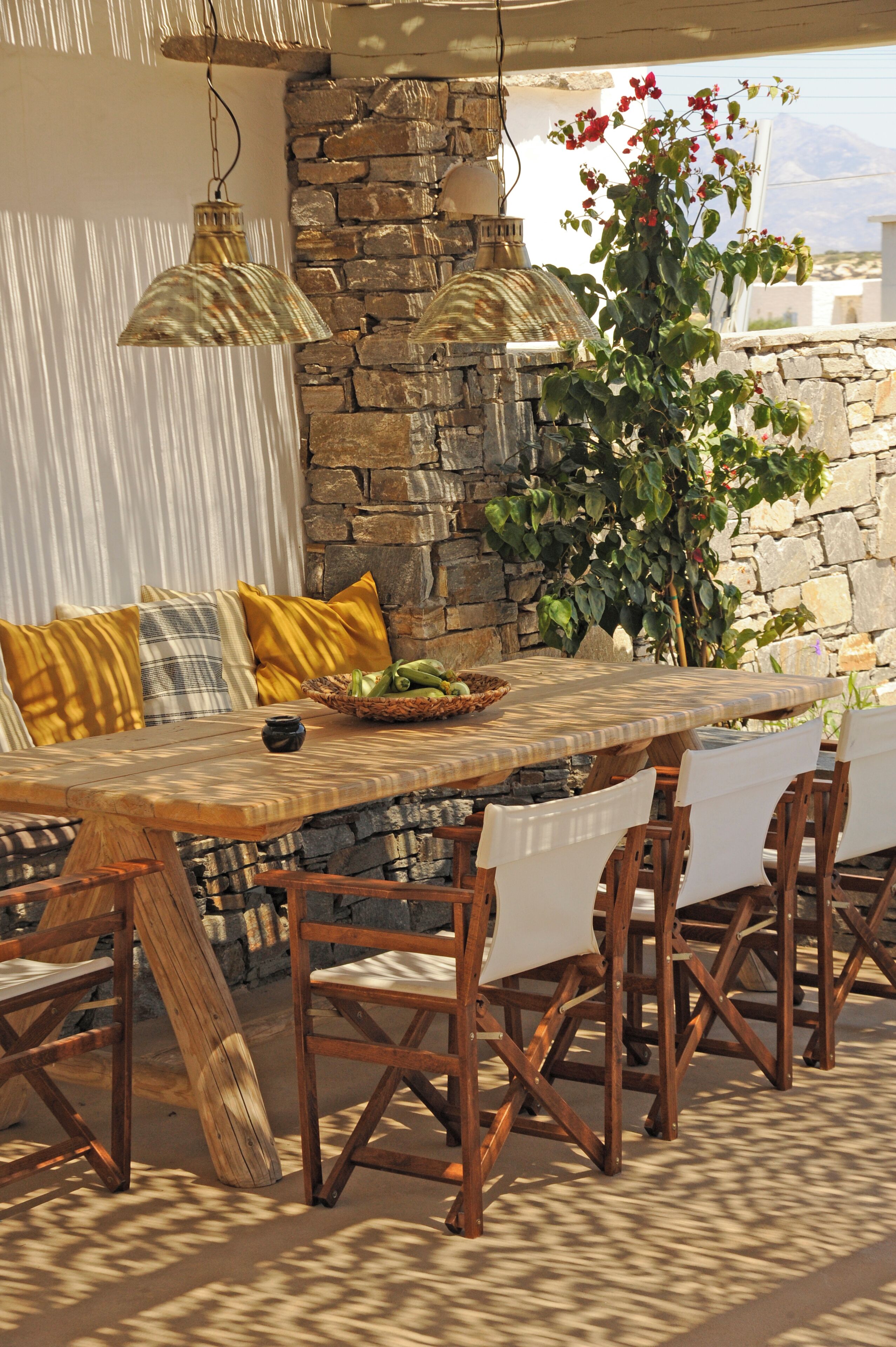 Uma mesa de jantar de madeira com cadeiras, uma parede de pedra, luminárias pendentes e um vaso com planta.