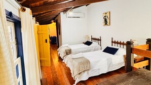 A room with wooden flooring, two beds with white linens, a wooden headboard, a small table, and a wall-mounted air conditioning unit. - Pousada Jardim Colonial (Tiradentes)