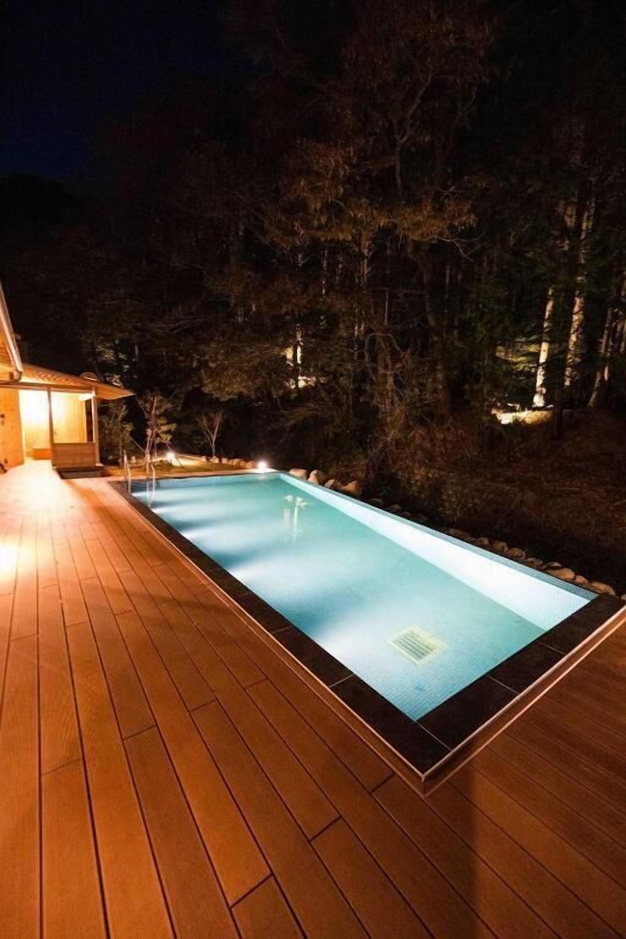 Una piscina rettangolare con un pontile in legno, circondata da alberi e un edificio illuminato.