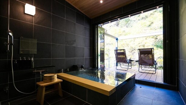 A modern bathroom with a large bathtub, a wooden stool, and a view of outdoor chairs through a glass door. - YOKOKAWAONSEN YUDONO TOTONOYU (Shimoda)