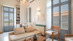 Una sala de estar con sofá, una silla de madera, una mesa con objetos decorativos, una estantería y vistas a través de un ventanal.