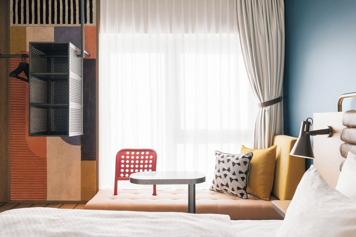 A hotel room with a bed, a small table, a red chair, and a window with curtains.