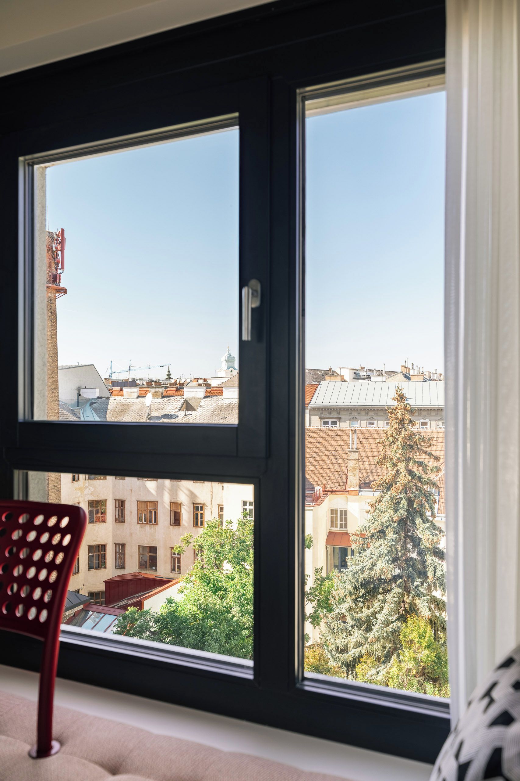 Une pièce dotée d’une grande fenêtre donnant sur un paysage urbain, d’un fauteuil rouge et d’un coussin à motifs.