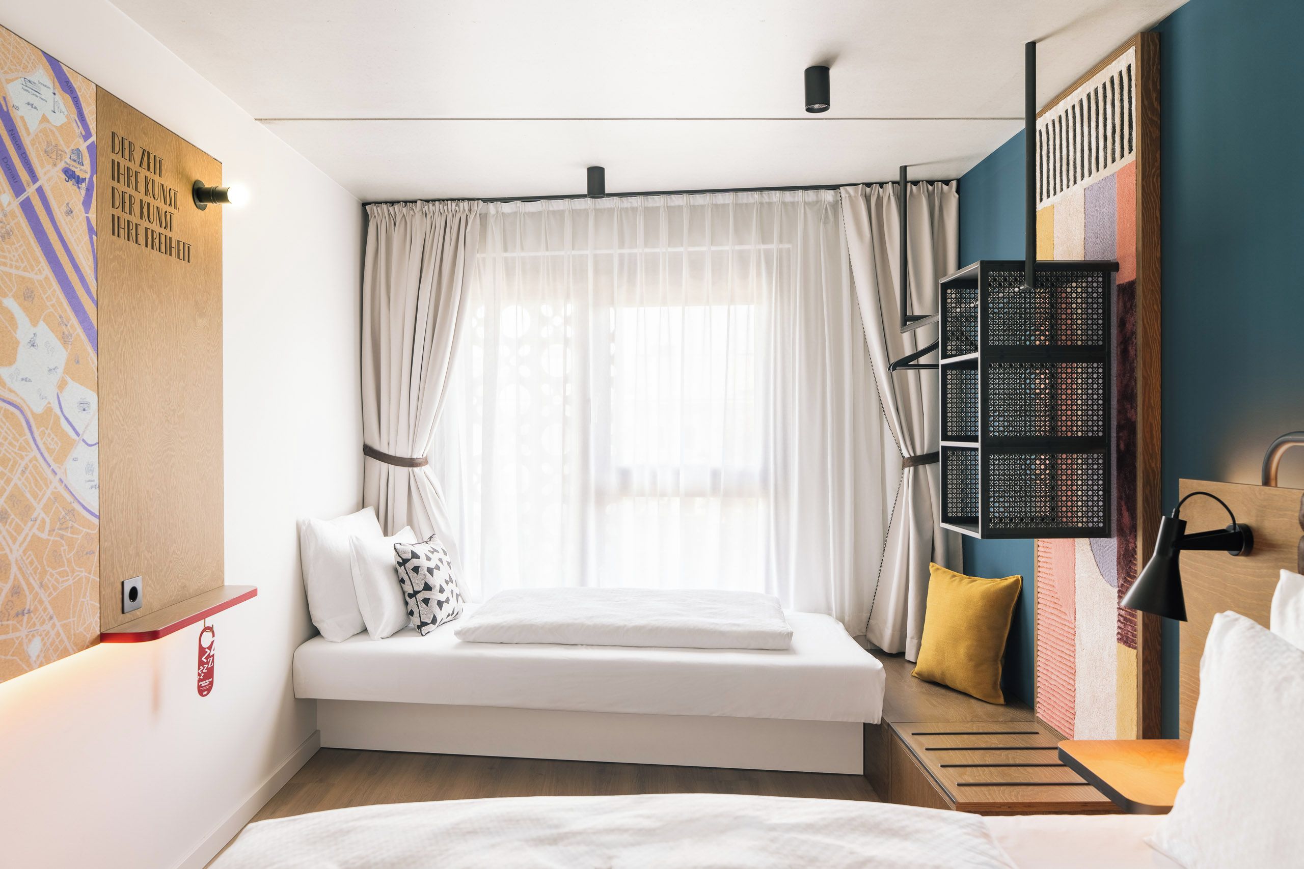 A modern hotel room with a bed, a map on the wall, a window with sheer curtains, and a small shelf with a lamp.