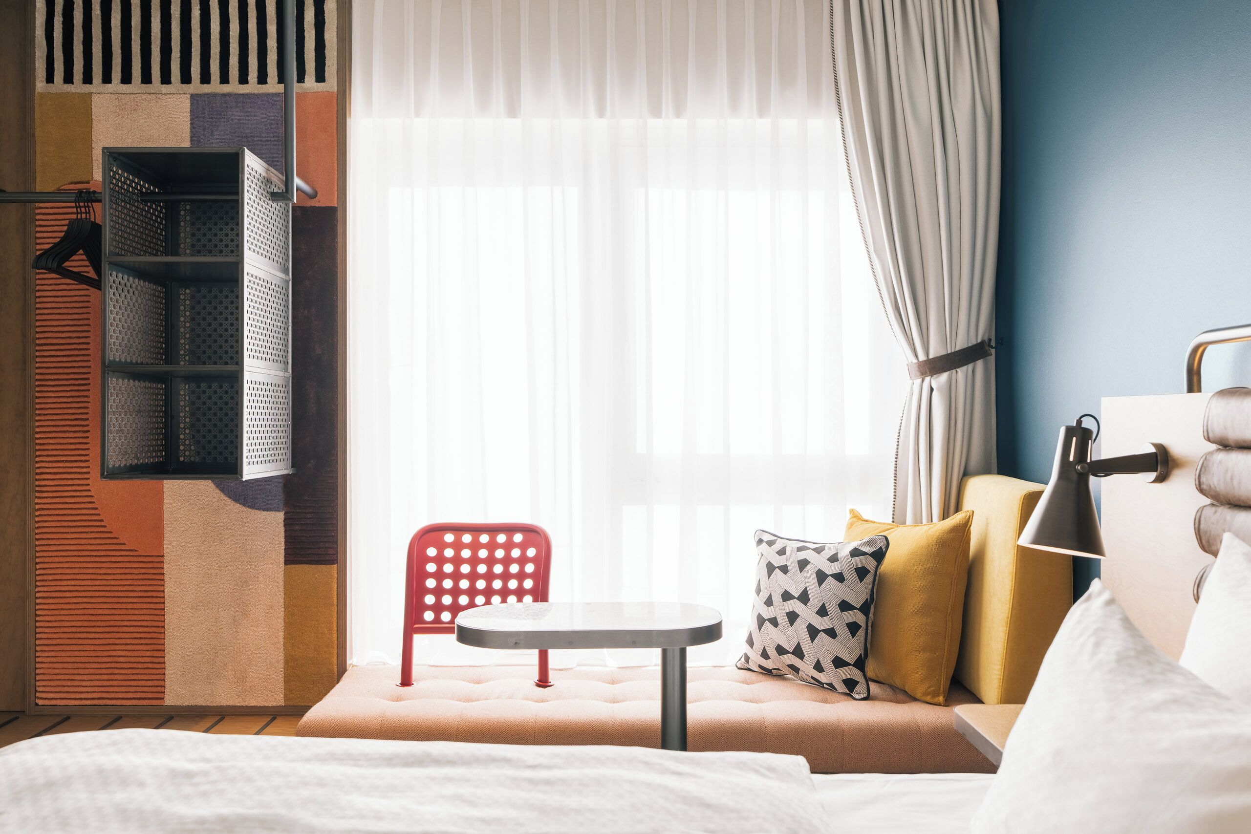 A hotel room with a bed, a small table, a red chair, and a window with curtains.