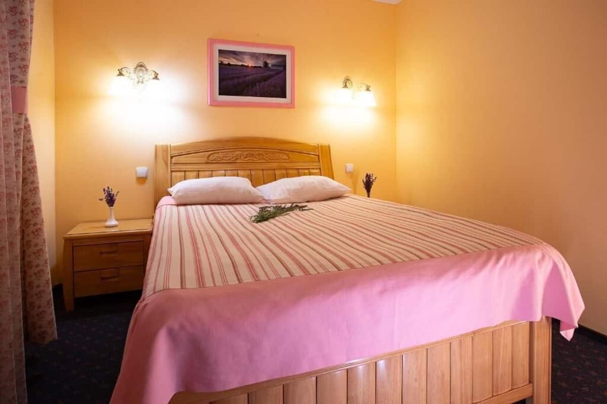 Una cama individual con cabecera de madera, dos lámparas de noche, una mesita de noche con lámpara y un cuadro en la pared.