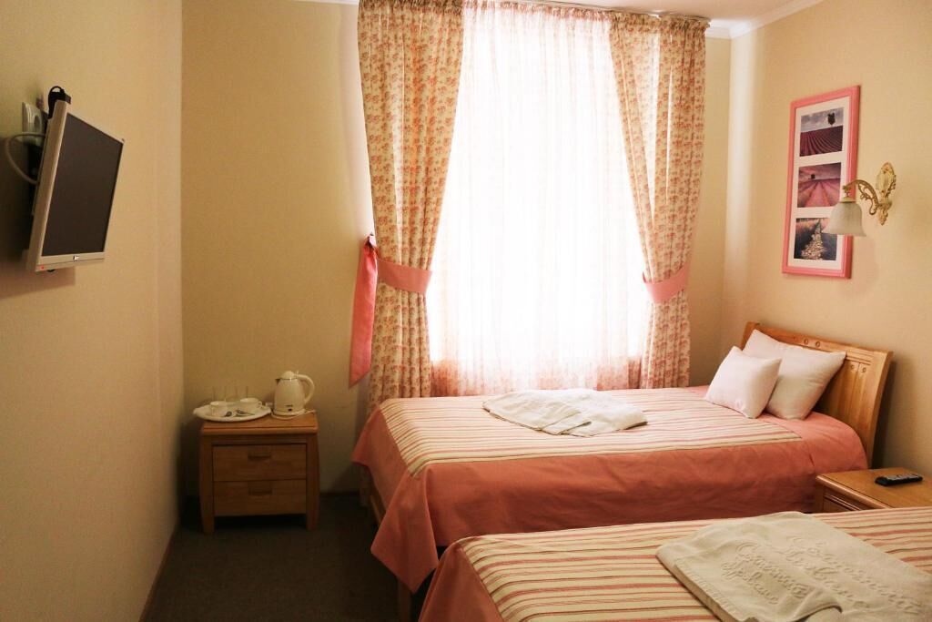 A hotel room with two beds, a bedside table with a kettle, a TV mounted on the wall, and a window with floral curtains.