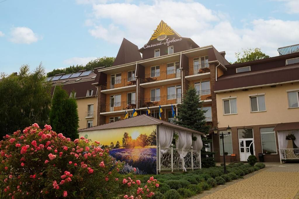 Un hotel de varios pisos con balcones, un jardín con flores rosadas y un mural en la fachada.