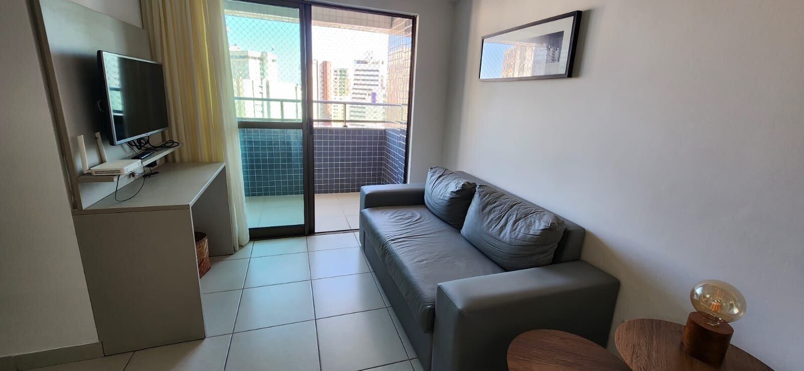 A modern living room with a flat-screen TV, a grey sofa, a small wooden table, and a balcony with a view of buildings.