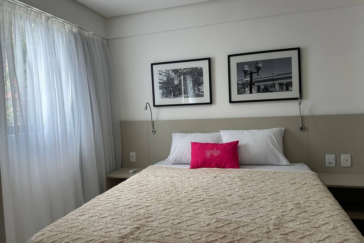 A hotel room with a bed, two framed pictures on the wall, a window with sheer curtains, and a bedside table with a remote control.