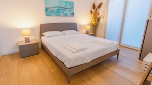 Una cama bien tendida con sábanas y toallas blancas, un cabecero gris y lámparas de noche.