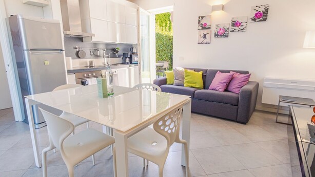 A modern kitchen with white cabinets, a dining table with chairs, and a grey sofa with colorful cushions. - Profumo di Rosa - One Bedroom Apartment, Sleeps 4 (Desenzano del Garda)