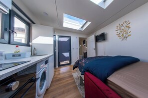 A compact room with a washing machine, a bed with a red and blue mattress, a small kitchen area with a sink and a window, and a skylight. - A Cozy Studio Apartment (Kilgobbin)