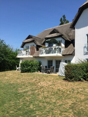 Ein zweigeschossiges Haus mit einem Strohdach, einem Balkon mit Gartenmöbeln und einer überdachten Terrasse.