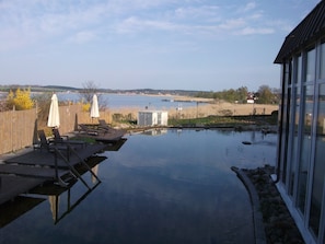 A swimming pool with lounge chairs and umbrellas, a wooden deck, and a view of a lake and distant buildings. - Apartment in Haus Wolgastsee with Shared Garden, Balcony, and Wi-Fi (Benz)