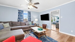 A living room with a sofa, a coffee table, a red chair, a ceiling fan, and a TV mounted on the wall. - NEW 4BR Home Nearby ACU & Downtown - Family Vacation - Summer Savings- Sleeps 10 (Abilene)