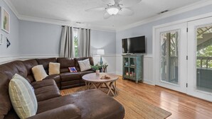 A living room with a brown leather sofa, a round wooden coffee table, a ceiling fan, a television on a stand, and a sliding glass door. - Sonesta III 202 by Seaside Vacations & Sales (Ocean City)