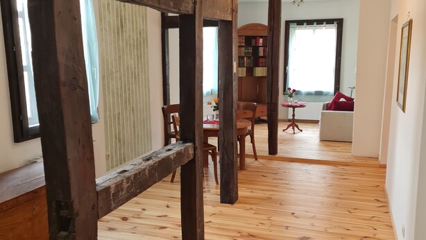 Ein geräumiger Raum mit Holzboden, sichtbaren Holzbalken, einer Essecke mit Stühlen und Tisch sowie einem Bücherregal.