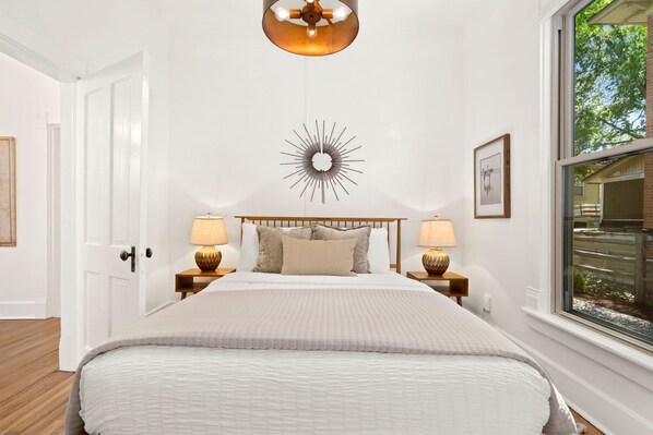 A bedroom with a bed, two bedside lamps, a wall clock, a window with a view, and a door.