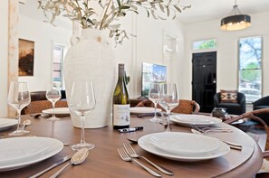 Uma mesa de jantar posta com pratos brancos, copos de vinho e talheres, uma garrafa de vinho ZUCCARDI e um vaso branco grande com galhos.