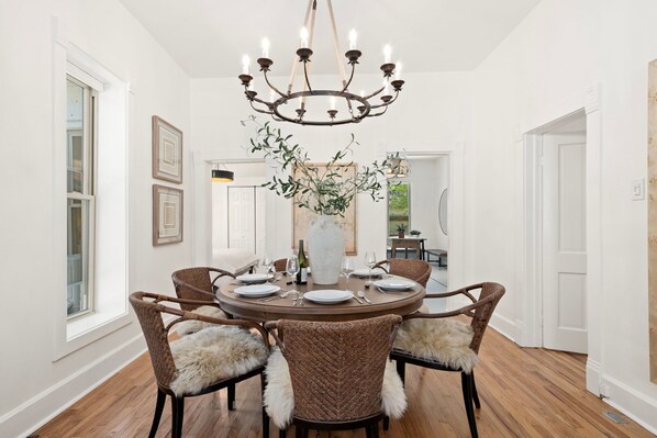 A dining room with a round table, wicker chairs with fur throw pillows, a chandelier, and a vase with greenery.