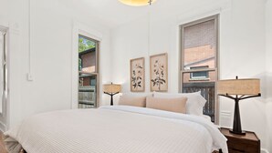 Una camera da letto con un letto grande, due comodini, una finestra vista su un edificio in mattoni e due opere d'arte appese al muro.