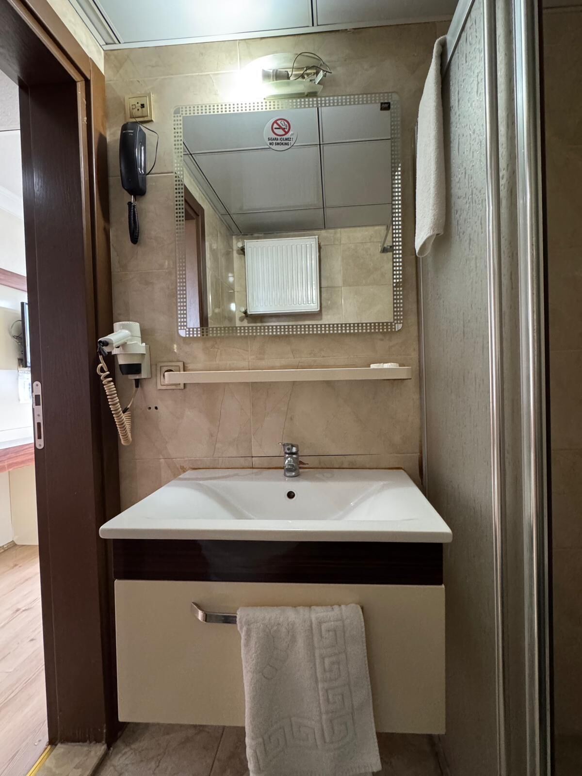 Un baño con un lavamanos blanco, un espejo, un perchero para toallas y un cartel de prohibido fumar.