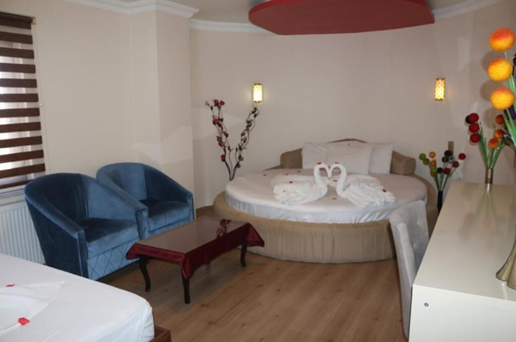A hotel room with a bed, two blue armchairs, a small table, a vase with flowers, and a wall-mounted light fixture.